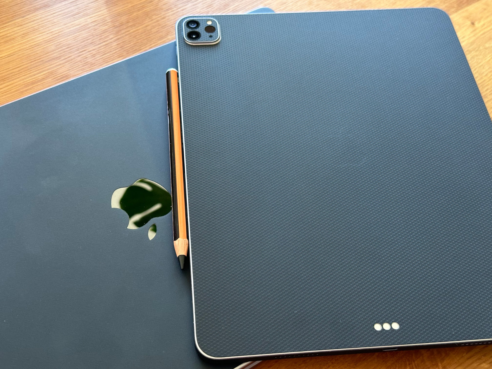 マットブラックのスキンが貼られたMacBook Pro 16インチの上に、マットブラック（Matrix）のスキンが貼られたiPad Pro 12.9インチが背面を上にして置かれている。iPadにはオレンジと黒のスキンでデコったApple Pencilがくっついている